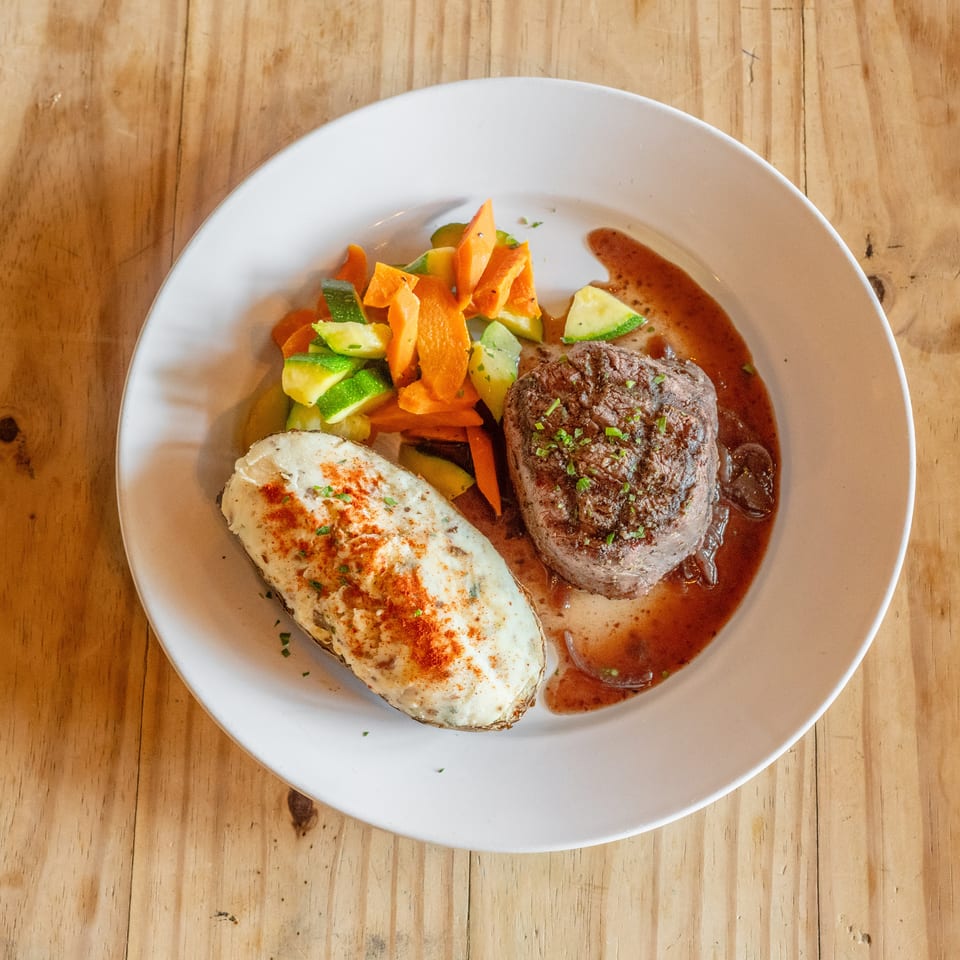 Grilled steak with creamy baked potato, roasted carrots and zucchini, served with brown sauce on a white plate.