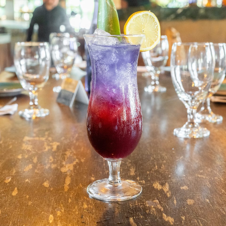 Layered purple and red cocktail with ice, lemon slice, and green garnish on a restaurant table surrounded by glassware.