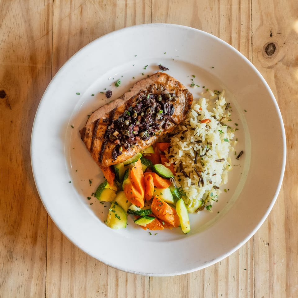 Grilled salmon topped with olive relish, wild rice, and roasted carrots and zucchini on a white plate.