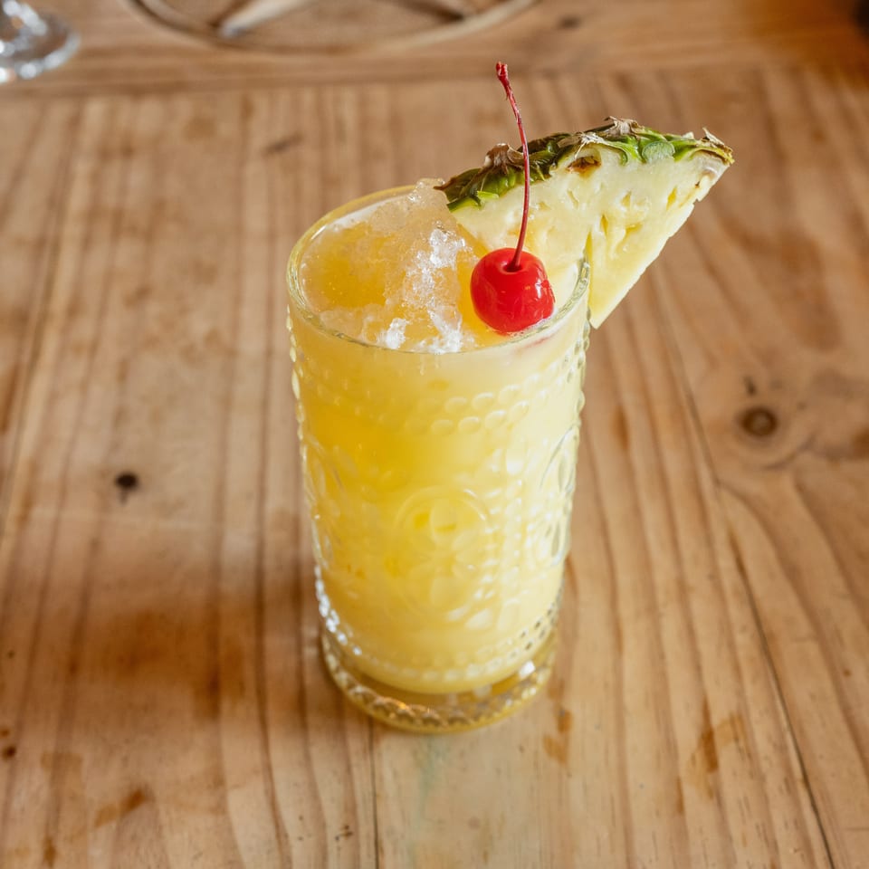 Yellow crushed-ice cocktail in a textured glass, garnished with a pineapple wedge and red cherry on a wooden table.