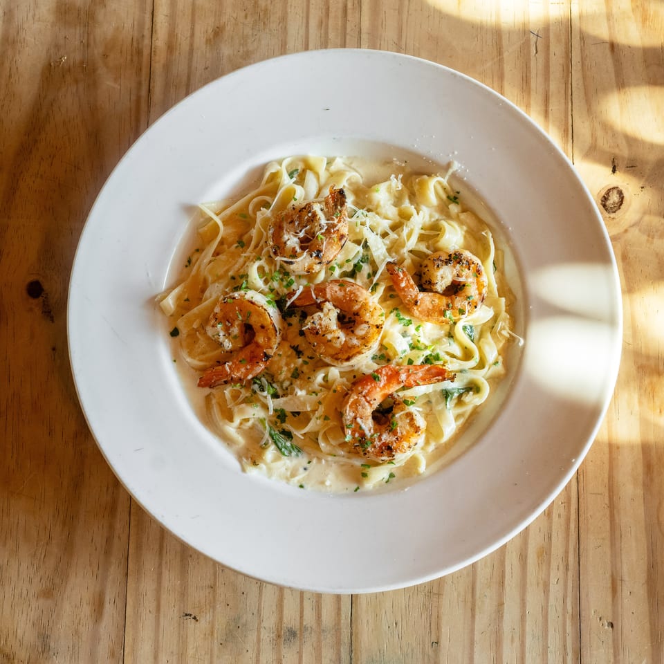 Grilled shrimp fettuccine Alfredo garnished with parsley and Parmesan in a white bowl on a wooden table.