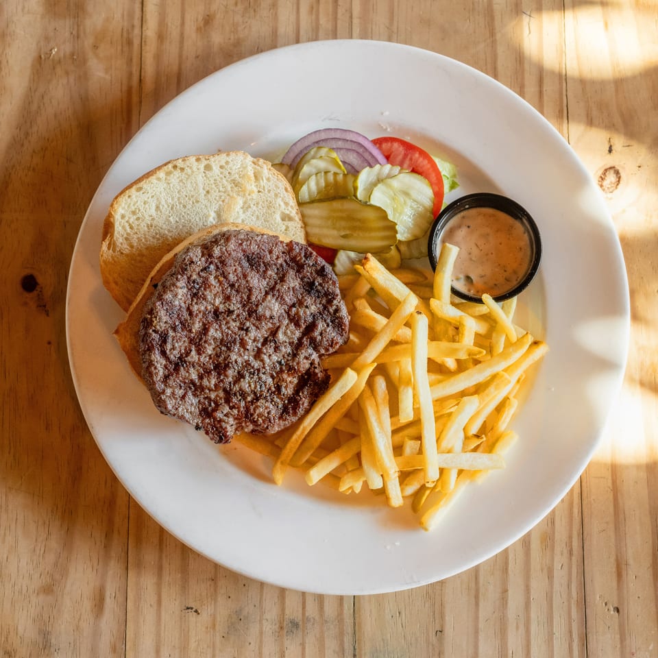 Grilled hamburger patty with bun, pickles, lettuce, tomato, onion, fries, and dipping sauce on a white plate.