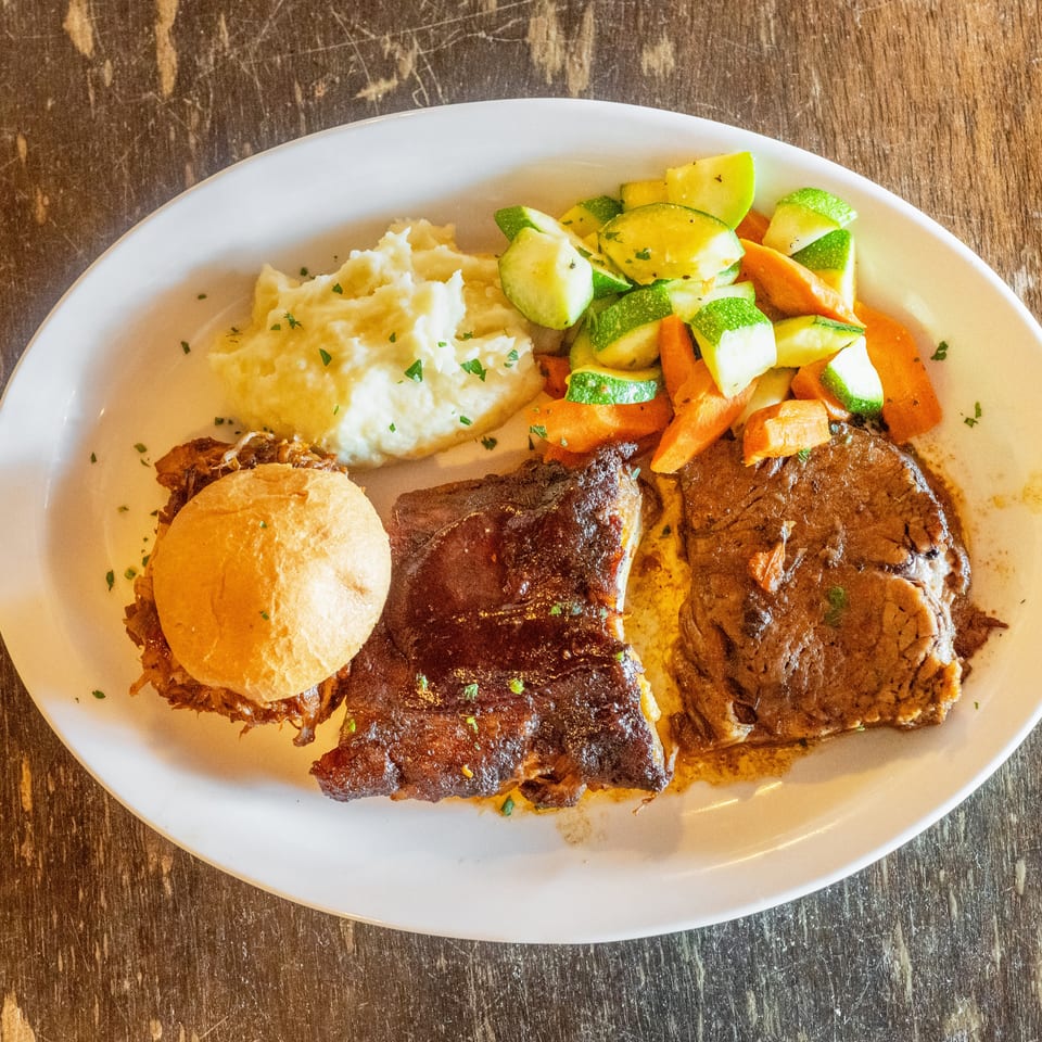 Plate of barbecue ribs, pulled pork sandwich, mashed potatoes, and roasted zucchini, squash, and carrots on a wooden table.