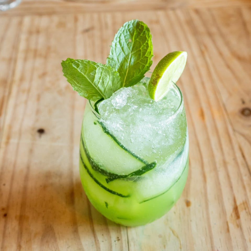 Refreshing lime and mint cocktail with crushed ice and cucumber slices in a glass on a wooden table.