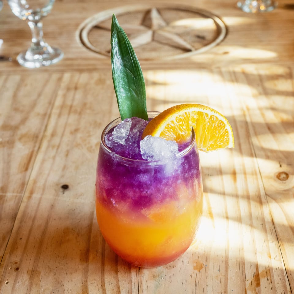 Colorful layered cocktail with purple ice, orange garnish, and green leaf on a sunlit wooden restaurant table.